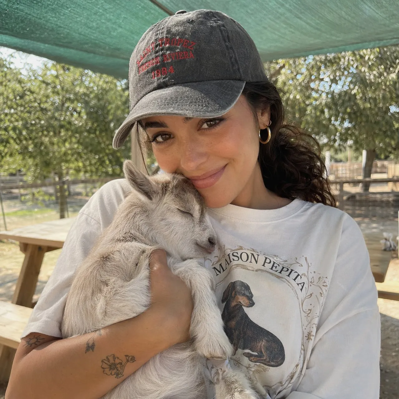 Close-up candid portrait of a young woman holding a baby goat, wearing oversized vintage graphic t-shirt and baseball cap, on a sunny outdoor farm
