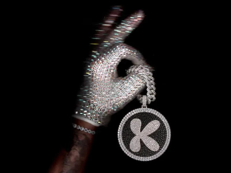 Round fully rhinestone-encrusted keychain with a rhinestone brand logo and a thick diamond chain threaded through the thumb of a sparkling crystal-gloved hand, white background, luxury product edit
