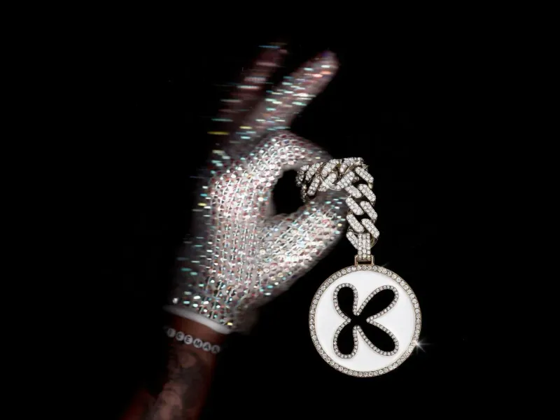 Round fully rhinestone-encrusted keychain with a rhinestone brand logo and a thick diamond chain threaded through the thumb of a sparkling crystal-gloved hand, white background, luxury product edit