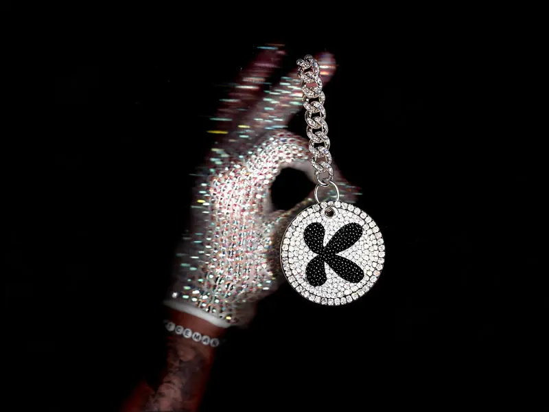 Round fully rhinestone-encrusted keychain with a rhinestone brand logo and a thick diamond chain threaded through the thumb of a sparkling crystal-gloved hand, white background, luxury product edit