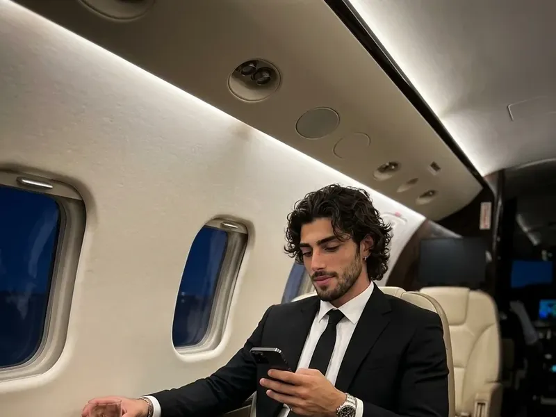 Photorealistic portrait of a man in a tailored black suit seated in a luxury private jet cabin at night, smartphone in hand, warm ambient lighting across cream and dark beige leather seating