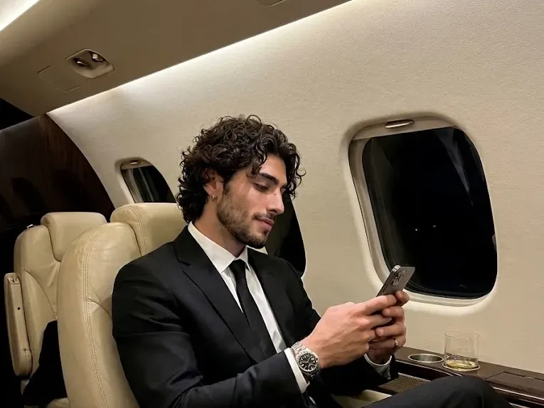 Photorealistic portrait of a man in a tailored black suit seated in a luxury private jet cabin at night, smartphone in hand, warm ambient lighting across cream and dark beige leather seating
