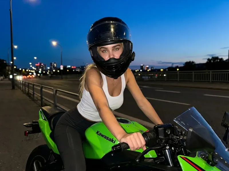 Photorealistic cinematic night street photo of a woman on a bright green Kawasaki-style sport motorcycle wearing a fitted tank top and partially open black helmet, with soft orange streetlight reflections under dusk twilight
