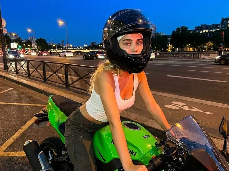 Photorealistic cinematic night street photo of a woman on a bright green Kawasaki-style sport motorcycle wearing a fitted tank top and partially open black helmet, with soft orange streetlight reflections under dusk twilight