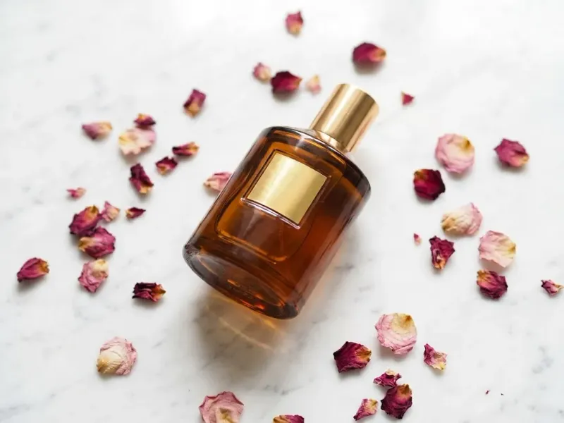 Perfume bottle on a white marble surface scattered with dried rose petals, soft diffused overhead lighting, editorial cosmetics product photography