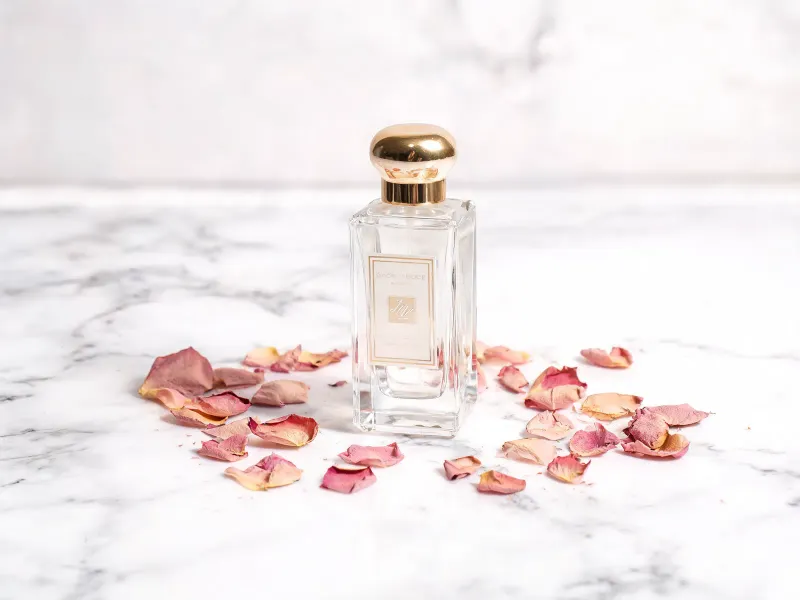 Perfume bottle on a white marble surface scattered with dried rose petals, soft diffused overhead lighting, editorial cosmetics product photography